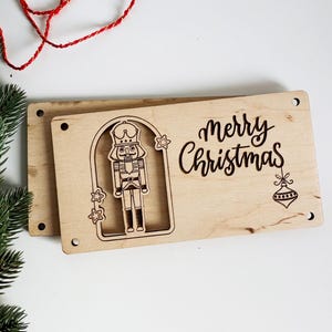 May include: Wooden Christmas sign with a laser-cut nutcracker design and the words "merry Christmas". The sign is rectangular with rounded corners and pre-drilled holes for hanging. A red string and green pine branch are in the background.