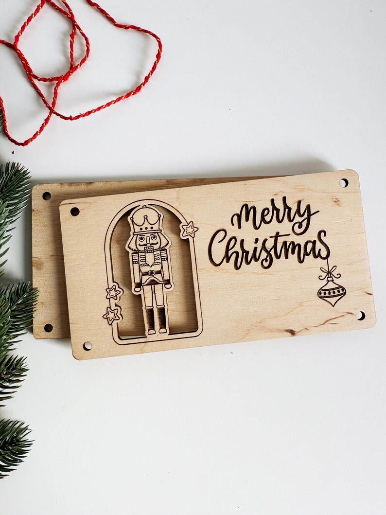 May include: Wooden Christmas sign with a laser-cut nutcracker design and the words "merry Christmas". The sign is rectangular with rounded corners and pre-drilled holes for hanging. A red string and green pine branch are in the background.