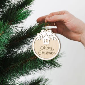 May include: A wooden Christmas bauble with a white cut-out design of a lantern and a bow. The bauble is hanging on a green Christmas tree and says "Merry Christmas" in cursive.