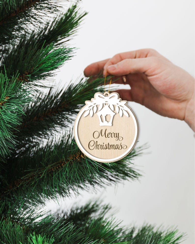 May include: A wooden Christmas bauble with a white cut-out design of a lantern and a bow. The bauble is hanging on a green Christmas tree and says "Merry Christmas" in cursive.