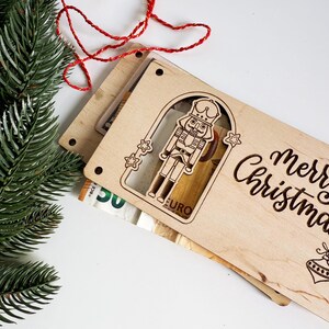 May include: Wooden Christmas gift card holder with a laser-cut nutcracker design and the words "Merry Christmas." The holder is made of light-colored wood and includes a red and gold string. Green pine branches are in the background.