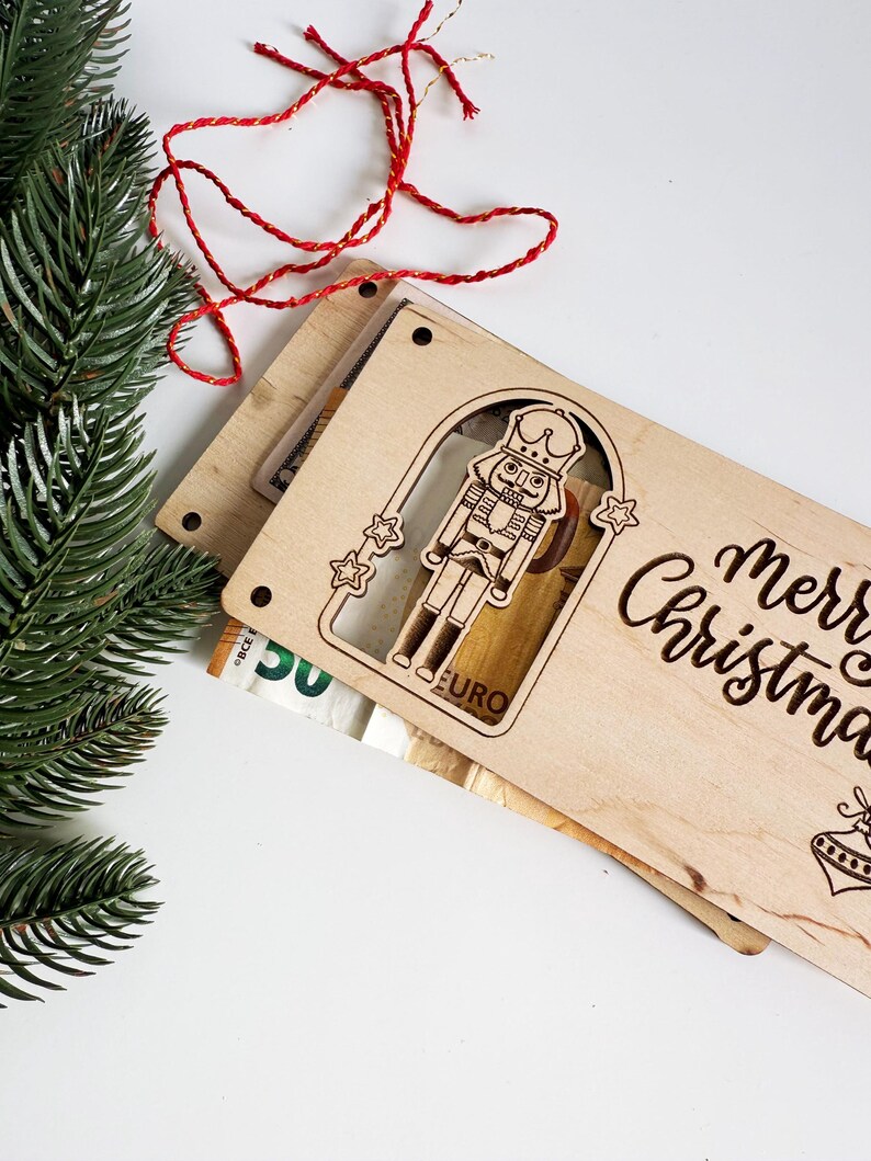 May include: Wooden Christmas gift card holder with a laser-cut nutcracker design and the words "Merry Christmas." The holder is made of light-colored wood and includes a red and gold string. Green pine branches are in the background.