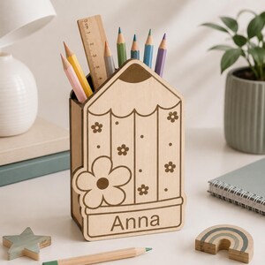 Puede incluir: Un portal&aacute;pices de madera de color claro con forma de l&aacute;piz grande, que presenta patrones de flores grabados y el nombre "Anna" en la parte frontal. Contiene una regla de madera y varios l&aacute;pices de colores. Tiene una base plana y parte superior abierta.