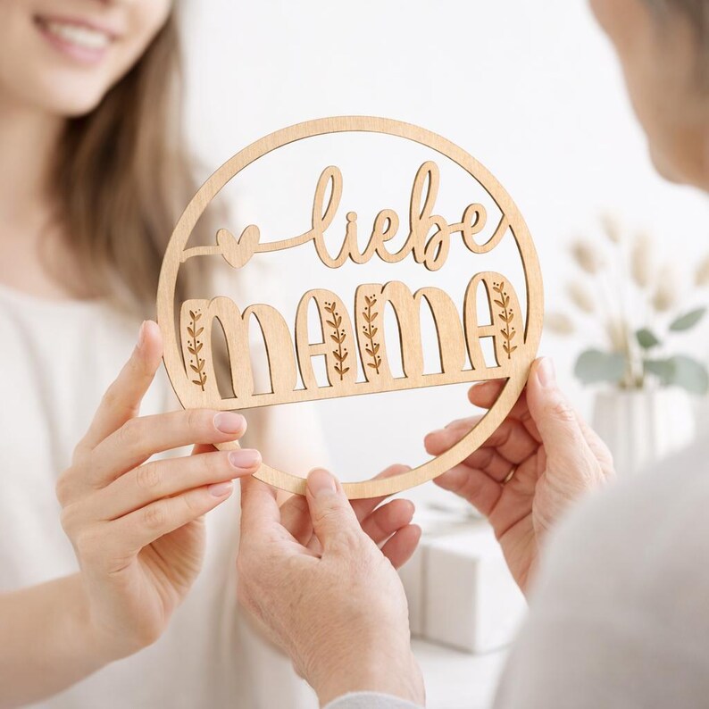 K&ouml;nnte beinhalten: Ein rundes Holzschild mit den Worten "liebe mama" in einer dekorativen Schriftart ausgeschnitten. Das Schild hat ein Herz und florale Akzente. Das Schild wird von zwei Personen gehalten.