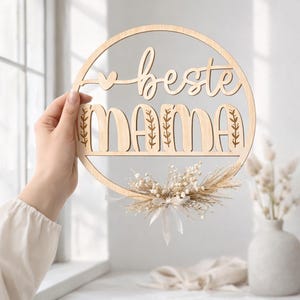 K&ouml;nnte beinhalten: Ein kreisf&ouml;rmiges Holzschild mit den Worten "beste mama" in einer Schreibschrift ausgeschnitten. Das Wort "mama" steht unter "beste" und hat florale Akzente. Ein kleiner Strau&szlig; aus Trockenblumen und Schleife ist unten befestigt.