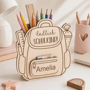Puede incluir: Un organizador de escritorio de madera clara con forma de mochila, con el texto Endlich SCHULKIND! y el nombre Amelia grabados en la parte frontal. El organizador contiene varios l&aacute;pices de colores y una regla de madera, con un bolsillo.