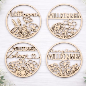 Pu&ograve; includere: Quattro cartelli circolari in legno tagliati al laser con disegni floreali e la parola "Willkommen" in vari stili. I cartelli sono marrone chiaro e presentano intricati dettagli ritagliati di fiori e fogliame.
