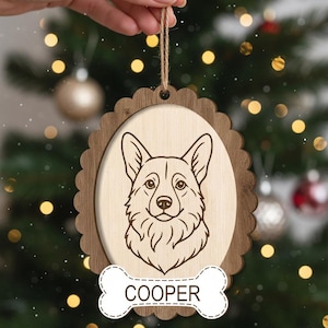 May include: Wooden Christmas ornament featuring a corgi dog portrait. The ornament has a light wood oval center with a detailed dog illustration, framed by a darker wood scalloped edge. A bone-shaped tag at the bottom reads "COOPER".