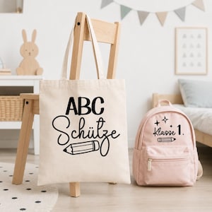 May include: A beige canvas tote bag featuring the black text ABC Sch&uuml;tze and a pencil graphic, displayed alongside a light pink backpack with the black text Klasse 1 and a pencil graphic with small star accents on the front pocket.