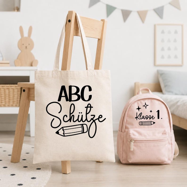 May include: A beige canvas tote bag featuring the black text ABC Sch&uuml;tze and a pencil graphic, displayed alongside a light pink backpack with the black text Klasse 1 and a pencil graphic with small star accents on the front pocket.