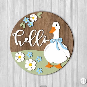 May include: Round wooden sign with a white goose wearing a blue bow, the word "hello" in white script, and white and blue flowers. The background is a wood grain pattern with a green and light green base.