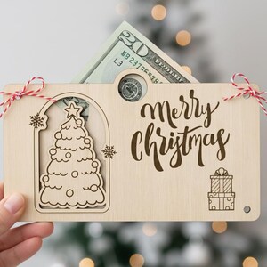 May include: A wooden Christmas gift card holder with a twenty-dollar bill peeking out. The holder features a laser-cut Christmas tree, snowflakes, and the words "Merry Christmas". It is tied with red and white twine.
