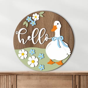 May include: A round wooden sign with a white "hello" script. A white goose with an orange beak and feet and a blue bow is on the right. The sign has a green field with white and blue flowers and a brown wood grain background.