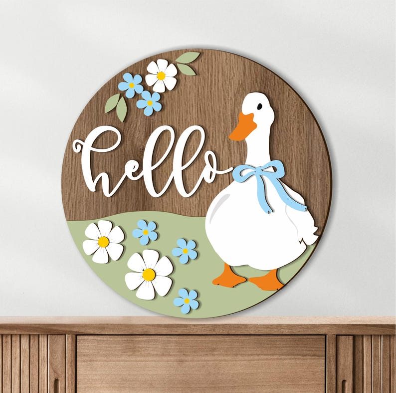 May include: A round wooden sign with a white "hello" script. A white goose with an orange beak and feet and a blue bow is on the right. The sign has a green field with white and blue flowers and a brown wood grain background.