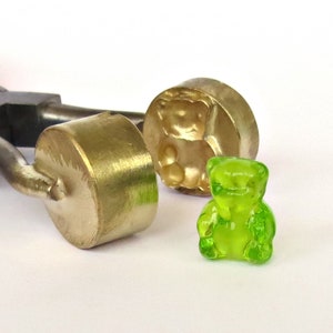 Könnte beinhalten: Ein Werkzeug mit rotem Griff und zwei Metallformen zum Herstellen von Gummibärchen. Eine Form ist ein Bärenmotiv, die andere ist rund. Ein grünes Gummibärchen liegt neben den Formen.