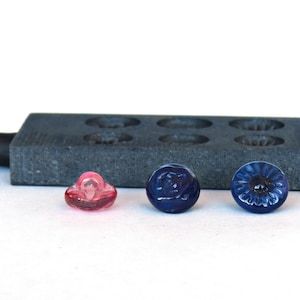 May include: Three glass buttons, one pink, one blue with a floral design, and one blue with a swirl design. The buttons are sitting on a grey surface.