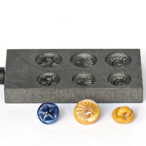 May include: A black metal mould with six circular indentations, each with a different design. Three glass buttons are shown below the mould: one blue with a star design, one brown with a shell design, and one brown with a swirl design.