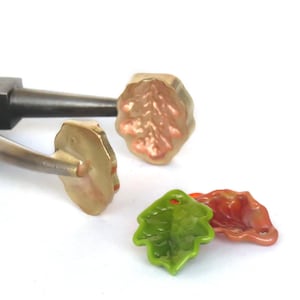 Könnte beinhalten: Nahaufnahme einer Zange, die einen goldenen, blattartigen Anhänger hält. Zwei weitere blattartige Anhänger liegen auf dem Tisch, einer grün und einer orange.