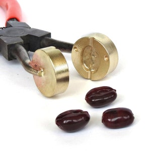 Puede incluir: Un par de alicates negros con mangos rojos sostienen dos moldes de latón. Los moldes tienen forma de granos de café. Tres granos de café rojo oscuro están sobre una superficie blanca.