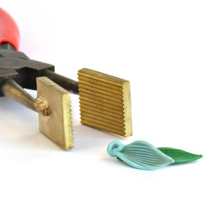 May include: A pair of pliers with two square metal attachments, one with a smooth surface and the other with a ridged surface. A light blue and green glass leaf pendant is in the foreground.