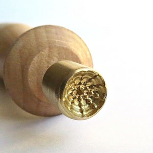 以下が含まれることがあります： 木製の麺棒で、金色の金属製の頭部には、テクスチャのある円形のデザインが施されています。頭部は木製のハンドルに取り付けられています。