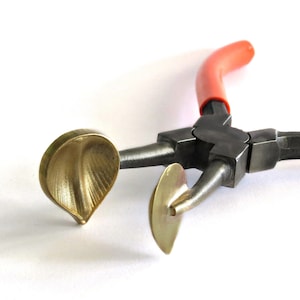 May include: A close-up shot of a jewellery-making tool. The tool has a black metal body with orange handles. The working ends are gold-coloured, leaf-shaped, and designed for shaping metal. The tool is set against a white background.