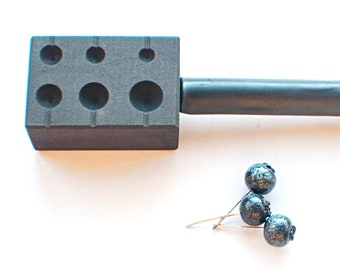 Può includere: Uno strumento in metallo nero con un manico e un blocco rettangolare con più fori rotondi. Tre perline blu scuro sono su una superficie bianca.