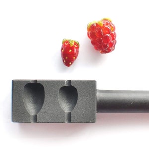 Op de afbeelding: Een zwarte metalen mal met twee holtes in de vorm van aardbeien, gebruikt voor het maken van glazen kralen. Twee rode glazen kralen, een in de vorm van een aardbei en een in de vorm van een framboos, worden naast de mal getoond.