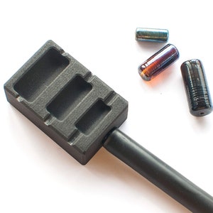 Op de afbeelding: Een zwarte metalen mal met drie rechthoekige holtes voor het gieten van glazen kralen. Drie glazen kralen, een blauwe, een bruine en een zwarte, liggen verspreid rond de mal.