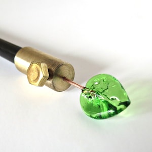 May include: A black and brass rod with a green glass leaf attached to the end. The leaf is shaped like a heart.