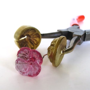 Puede incluir: Un par de alicates con un mango rojo sostiene dos moldes de latón para hacer flores de vidrio. Una flor de vidrio rosa está sobre la superficie blanca.