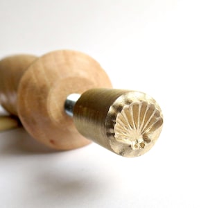 Puede incluir: Un mango de madera con un sello de latón que presenta un diseño detallado de una concha. El sello se utiliza para crear impresiones en arcilla o masa.