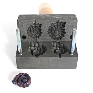 Könnte beinhalten: Eine graue Metallform mit vier Hohlräumen in Form von Ammoniten. Die Form ist geöffnet und ein lila Ammonit liegt davor.