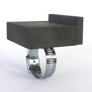Op de afbeelding: Een grijze betonnen blok met een verhoogde rand, bevestigd aan een zilveren metalen klem.