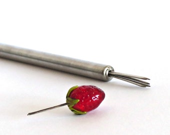 Può includere: Un attrezzo per togliere il torsolo delle fragole in acciaio inossidabile con una fragola rossa, simile al vetro. L'attrezzo ha più rebbi a un'estremità. La fragola ha foglie verdi e un gambo.