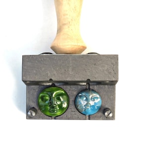 Op de afbeelding: Een metalen mal met twee cirkelvormige holtes, elk met een glazen gezichtshanger. De ene hanger is groen, de andere blauw met zilveren accenten. Een houten handvat is aan de mal bevestigd.