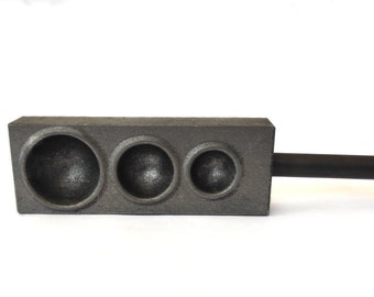 May include: A dark gray metal casting mold with three circular depressions of varying sizes. A black handle extends from the side. The mold is designed for shaping materials, likely in a crafting or jewelry-making context.