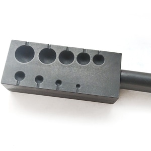 May include: A black metal tool with a handle and a rectangular block with multiple round depressions of varying sizes. The tool is used for shaping metal.
