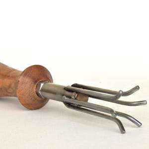 Puede incluir: Una herramienta para trabajar la madera con un mango de madera y una cabeza de metal con múltiples púas curvas. El mango es de color marrón claro y los componentes metálicos son plateados.