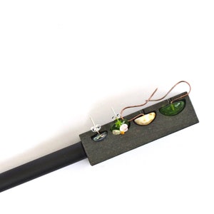 Op de afbeelding: Drie paar oorbellen die op een zwarte displaystandaard worden getoond. De oorbellen zijn gemaakt van glas en hebben groene, witte en gele kleuren.