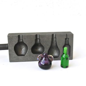 Puede incluir: Un molde gris oscuro con cuatro cavidades en forma de botella, junto a un pequeño jarrón de cristal morado con asas y una botella de cristal verde. El molde tiene un mango largo. Las botellas son de diferentes formas.