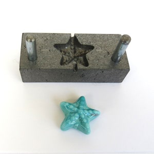 May include: A grey concrete mould with two metal rods and a star-shaped cavity. A teal blue glass star is sitting next to the mould.