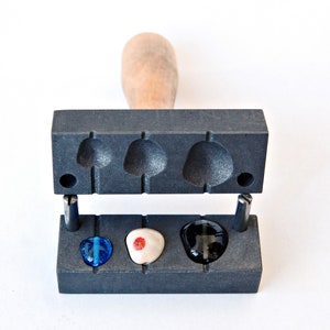 Op de afbeelding: Een zwarte metalen mal met vier holtes voor het maken van glazen cabochons. De mal is open en toont drie cabochons: een blauwe, een witte met een rode bloem en een zwarte.