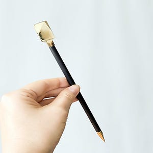 Puede incluir: Una herramienta larga y delgada con un eje cilíndrico negro mate, una cabeza metálica de tono dorado pulido con borde plano en la parte superior y una punta metálica de tono dorado en el extremo inferior.