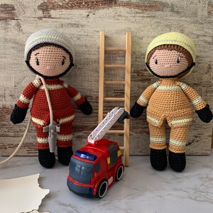 Könnte beinhalten: Zwei gehäkelte Feuerwehrpuppen, eine in Rot und eine in Braun, stehen neben einer Holzleiter und einem roten Spielzeugfeuerwehrauto mit einer Leiter.