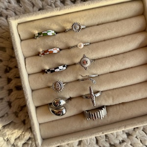 Könnte beinhalten: Ein Set aus neun silbernen Ringen in einer weißen Box. Die Ringe haben verschiedene Designs, darunter eine Blume, eine Schlange, ein Kreuz und geometrische Muster. Einige Ringe sind mit buntem Email verziert.