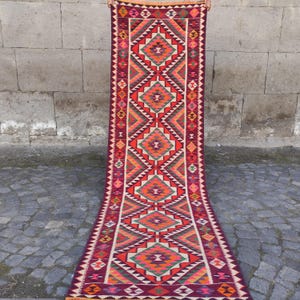 May include: A long, rectangular rug with a geometric pattern. The rug features a deep burgundy background with vibrant diamond and triangle shapes in red, orange, green, and cream. The edges are finished with a fringe.