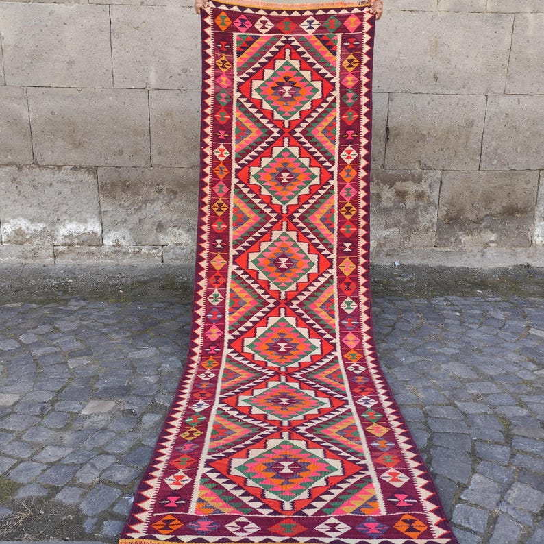 May include: A long, rectangular rug with a geometric pattern. The rug features a deep burgundy background with vibrant diamond and triangle shapes in red, orange, green, and cream. The edges are finished with a fringe.