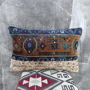 May include: Rectangular decorative pillow with a brown, blue, and beige patterned design. The pillow features a central brown section with intricate geometric patterns in blue, purple, and green. The top and bottom borders are blue and beige, respectively. The pillow is placed on a patterned surface.