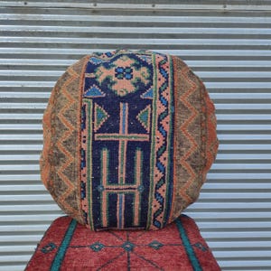 Puede incluir: Cojín redondo estampado con un diseño geométrico central en azul marino, turquesa y melocotón. El cojín tiene un borde marrón con patrones geométricos naranjas y beige. Descansa sobre una alfombra roja con detalles turquesa.